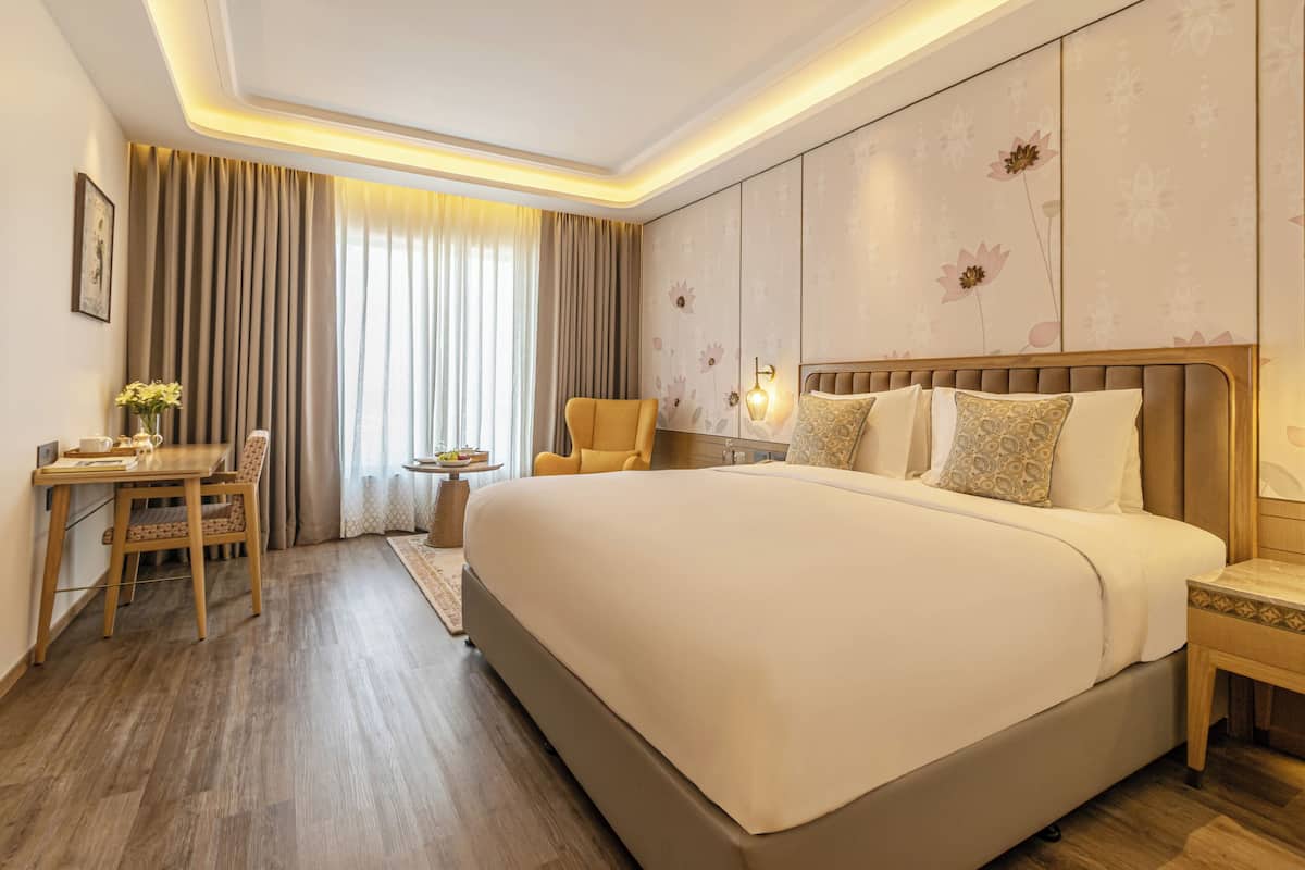 Habitación de hotel con una cama grande, un escritorio con silla, una mesita de noche con lámpara y un ventanal con cortinas.