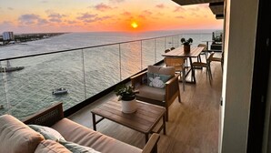 Ein Balkon mit einer Couch, Sesseln und einem Tisch mit Blick auf einen Sonnenuntergang über dem Wasser.