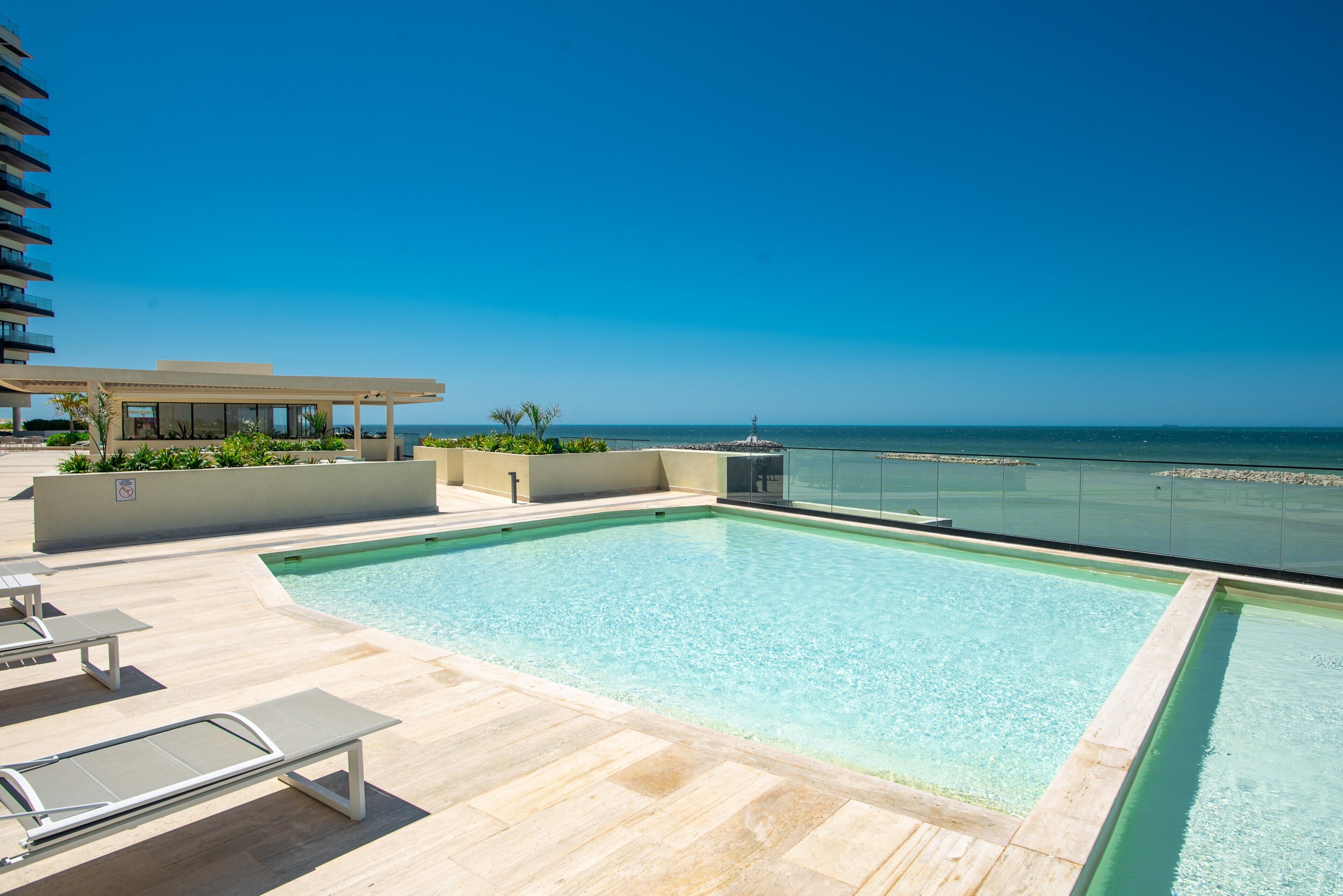 A rooftop pool with a clear view of the ocean, lounge chairs, and a building with a balcony.