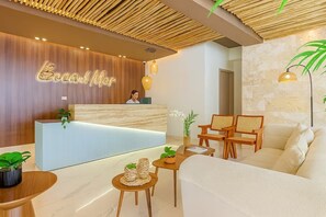 Un área de recepción con un mostrador de madera, una recepcionista, un sofá, dos sillones y una mesita con plantas.