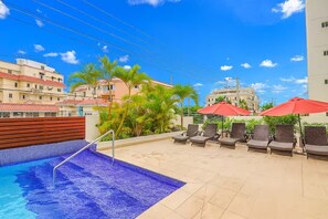Una piscina en la azotea con sillones de descanso y sombrillas, rodeada de edificios y palmeras.