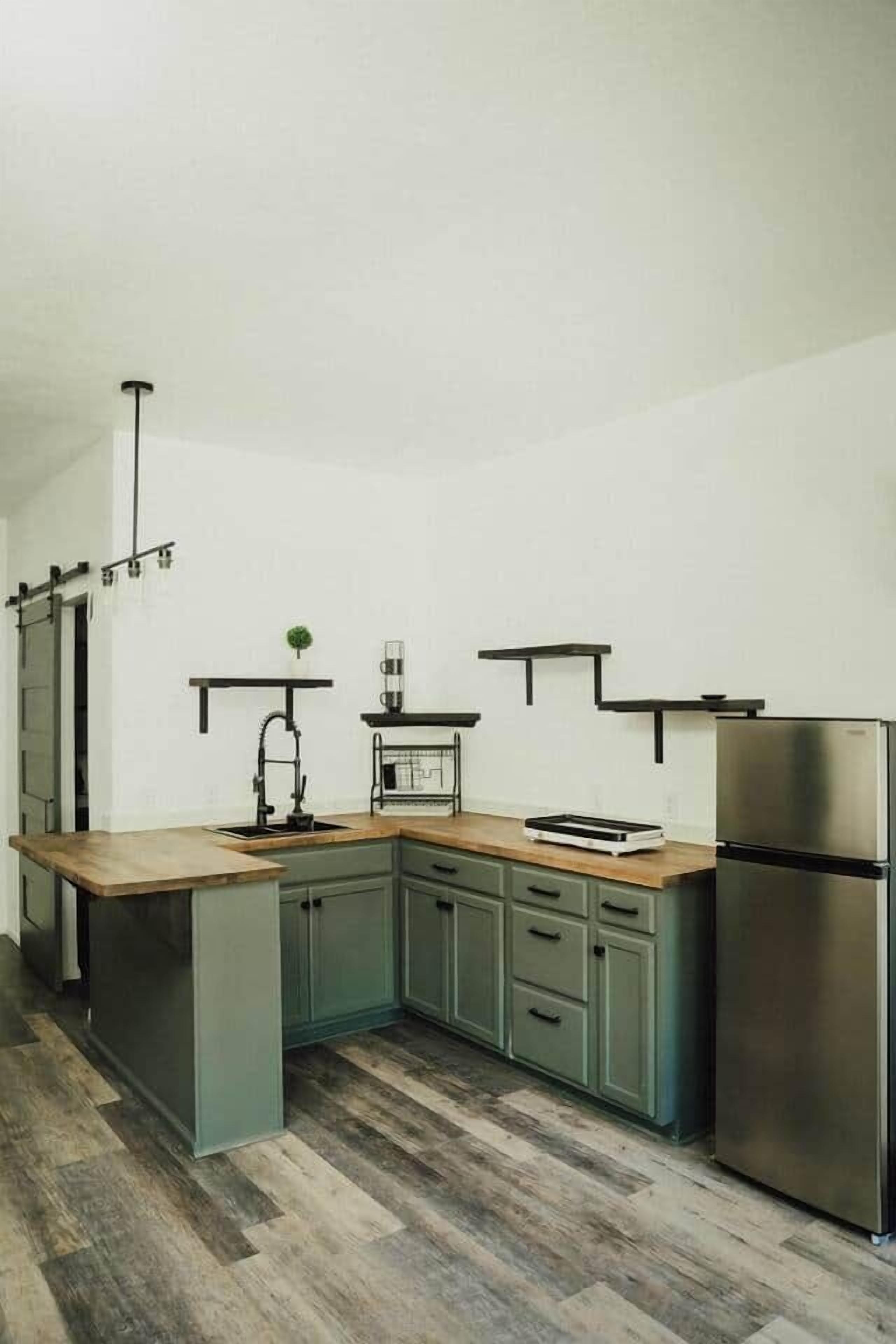 A modern kitchen with a wooden countertop, green cabinets, a stainless steel refrigerator, and a hanging light fixture.