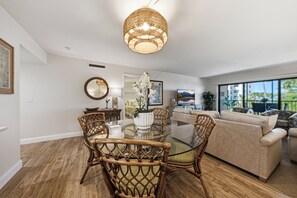 Un salon moderne avec une table à manger en verre, des chaises en osier, un canapé et une grande fenêtre donnant sur l’extérieur.