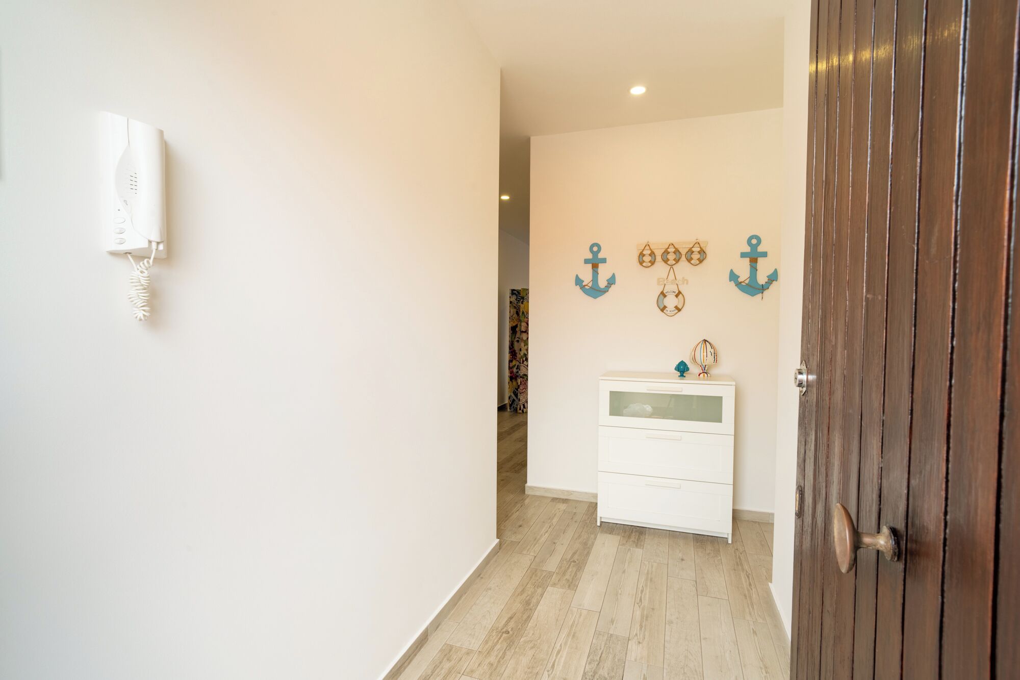 Un corridoio con una porta in legno, un telefono bianco fissato al muro e un comò bianco con cassetti.