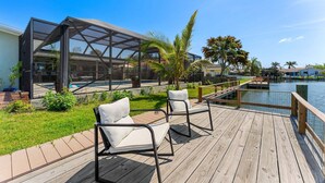A wooden deck with two white cushioned chairs, a glass-enclosed structure, and a pool. - Tropical Cocoa Beach Riverfront Oasis | Pool, Hot Tub & Lanai 🍹🌴 (Cocoa Beach)