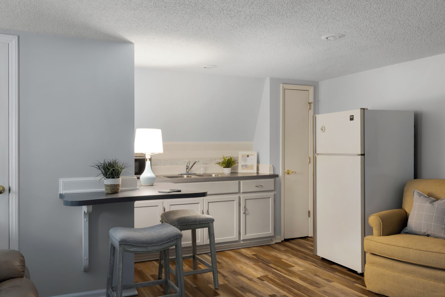 A compact kitchen with a countertop, sink, cabinets, and a refrigerator. There are two stools and a chair.