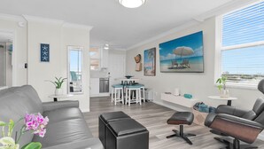 Un salon moderne avec un canapé gris, un pouf noir et un fauteuil en cuir noir. On y trouve également un coin cuisine avec des meubles blancs et une table à manger entourée de chaises bleues. Un tableau représentant une scène de plage est accroché au mur.