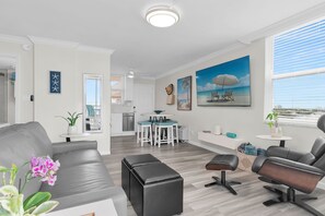 Ein modernes Wohnzimmer mit grauem Sofa, schwarzem Hocker und einem schwarzen Ledersessel. Ein Küchenbereich mit weißen Schränken und einem Esstisch mit blauen Stühlen. Auf der Wand hängt ein Gemälde, das eine Strandlandschaft zeigt.
