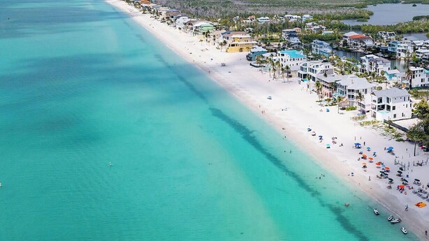 Vue aérienne d’une zone côtière avec une longue plage de sable, des eaux turquoise et limpides, et une zone résidentielle densément construite, avec des bâtiments situés près du rivage.