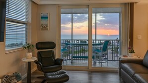 Un salon avec vue sur le coucher de soleil sur l’eau, un fauteuil en cuir noir, une petite table avec une plante et un canapé.