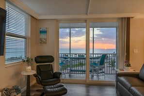 Ein Wohnzimmer mit Blick auf den Sonnenuntergang über dem Wasser, ein schwarzer Ledersessel, ein kleiner Tisch mit einer Pflanze und ein Sofa.