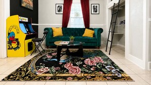 Un salon avec un canapé vert, une table basse, un tapis à motifs floraux, une borne d’arcade jaune et une échelle dans un coin.