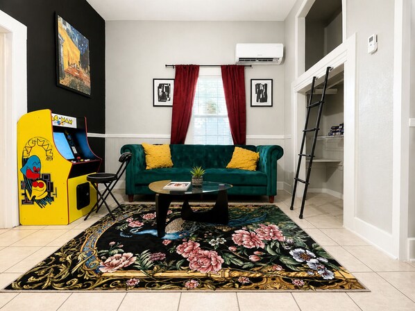 A living room with a green sofa, a coffee table, a floral rug, a yellow arcade machine, and a ladder in the corner.