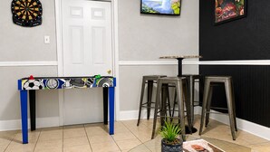 Une pièce comprenant un jeu de fléchettes, un téléviseur, un tableau encadré, une table avec des tabourets et une plante en pot.