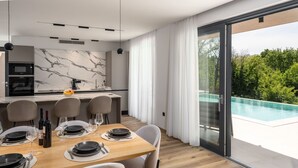Uma área de jantar moderna com mesa de madeira posta para seis, cozinha com painel de fundo em mármore e porta de vidro que dá para uma piscina.