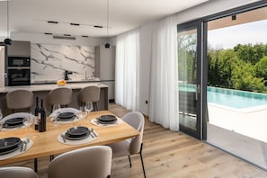 A modern dining area with a wooden table set for six, a kitchen with marble backsplash, and a glass door leading to a pool. - Villa Vanja (Istria - Poreč) (Poreč)