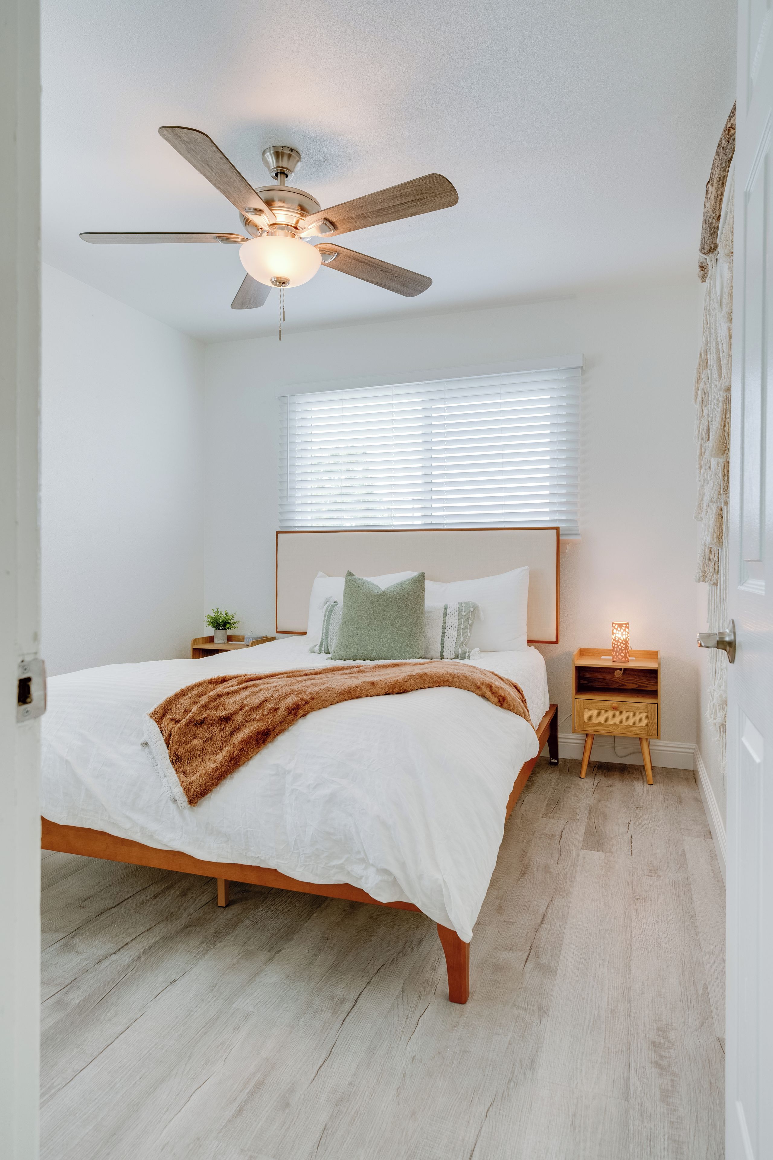 Un dormitorio con cama, mesita de noche, lámpara de pared, ventilador de techo y ventana con persianas.