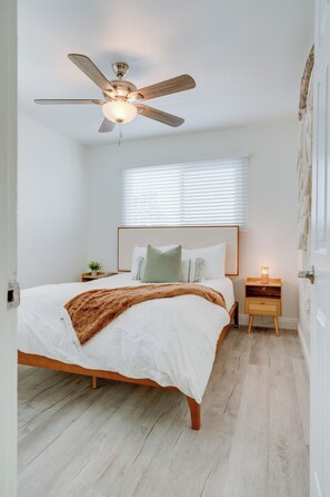 A bedroom with a bed, a nightstand, a wall-mounted lamp, a ceiling fan, and a window with blinds.