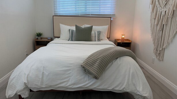 Una cama bien hecha con edredón blanco y almohadas, una lámpara de noche y un tapiz de macramé en la pared.