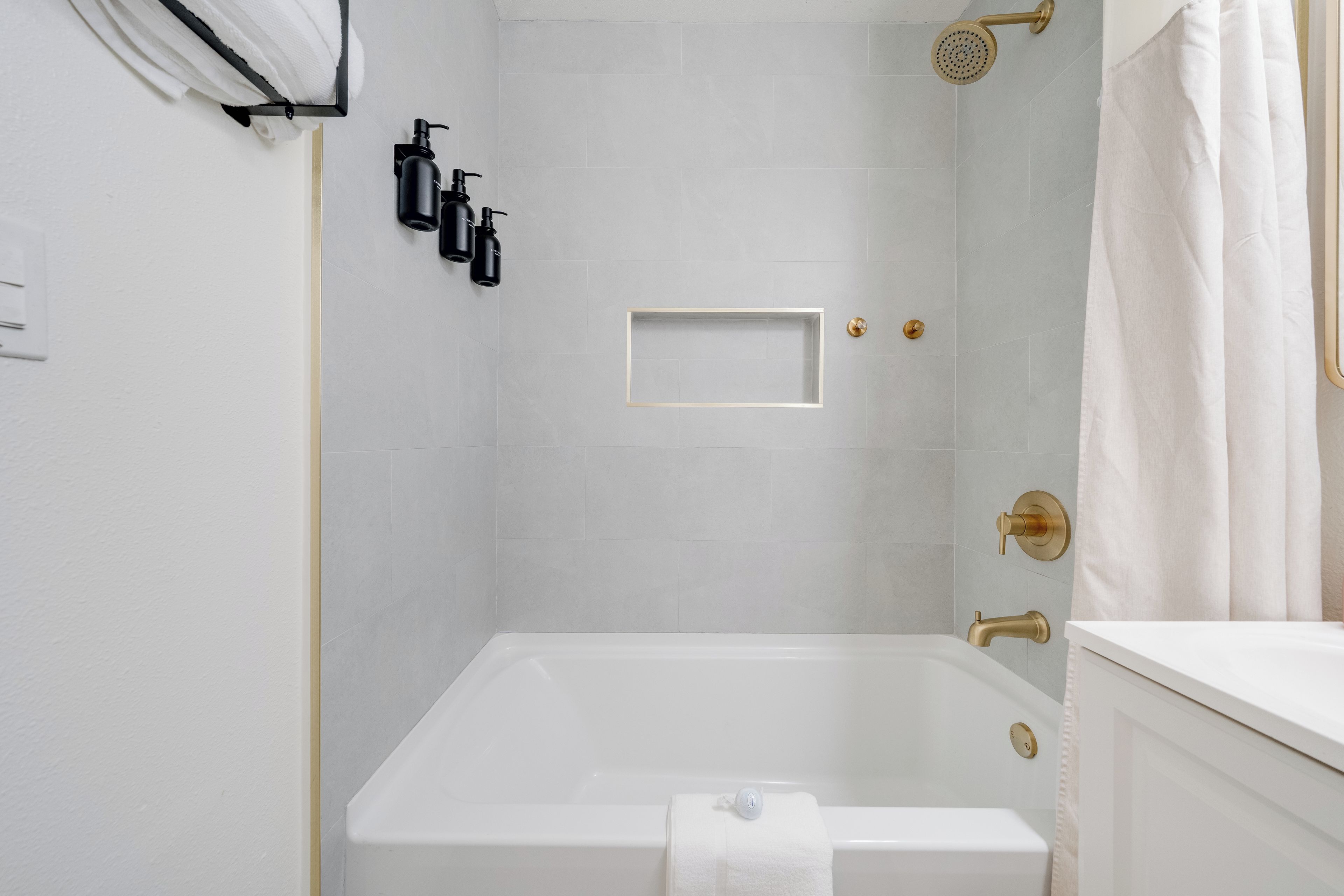 Ein modernes Badezimmer mit einer weißen Badewanne, einer Dusche mit goldfarbener Duschbrause und einem weißen Handtuch an der Wand.