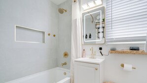 Un baño moderno con una bañera, ducha, tocador con espejo y ventana con persianas.