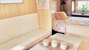 A cozy interior with a sofa, a small table with decorative items, a bed with a floral bedspread, and a window with a view. - Elizabeth the spacious retro caravan. Sleeps 6 | Rye, Mornington Peninsula (Rye)