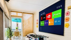 Een kamer met een tafelvoetbaltafel, een bank, een groot scherm met app-icoontjes en een raam met luxaflex.