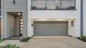 A two-story house with a grey garage door, a balcony with a metal railing, and a small garden area with plants.