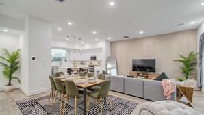 A modern living room with a dining area, a grey sofa, a TV, and a kitchen in the background.