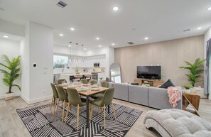 Ein modernes Wohnzimmer mit Essbereich, einem grauen Sofa, einem Fernseher und einer Küche im Hintergrund.
