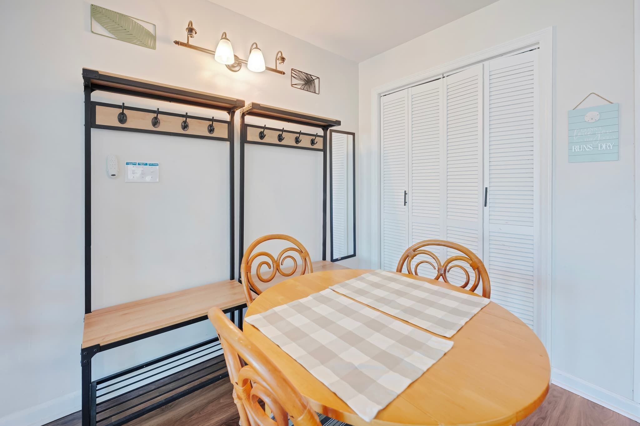 Ein Zimmer mit einem Holzespeisetisch, zwei Stühlen, einer Bank, einem Kleiderhaken mit Haken, einer wandmontierten Leuchte, einer Tür mit Klappläden und einer kleinen Wanddekoration.