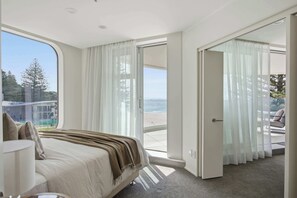 A bedroom with a bed, a nightstand, a lamp, and a view of the beach through the window. - Marine Horizon Beachfront, Pool, Spa, Sauna & Gym (Mount Maunganui)