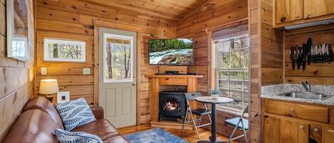 A log cabin interior with a fireplace, a television, a sofa, and a kitchen area.