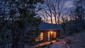 A log cabin with a porch, surrounded by trees and a gravel path.