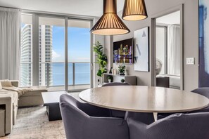 Un salon moderne avec une table à manger ronde, des chaises recouvertes d’un tissu gris, un canapé et une grande fenêtre offrant une vue sur les immeubles de grande hauteur et la mer.