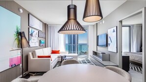 Ein modernes Hotelzimmer mit einem großen Esstisch, einer Couch mit orangefarbenen Kissen, einem Fernseher und einem Balkon mit Blick auf die Stadt.