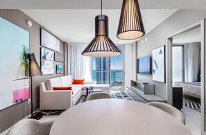Une chambre d’hôtel moderne avec une grande table à manger, un canapé avec des coussins orange, une télévision et un balcon offrant une vue sur la ville.