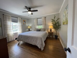 Ein Schlafzimmer mit einem Bett, zwei Nachttischen, einem Deckenventilator, einem Fenster mit Vorhängen, einer Topfpflanze und einer Tür mit Knauf.