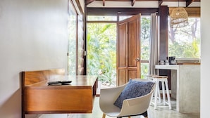 Una habitación con un escritorio de madera, una silla blanca con cojín azul, una zona de cocina y un amplio ventanal que da a un jardín.