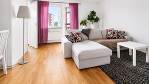Una sala de estar moderna con sofá, reposapiés y mesa de centro, una lámpara de pie y vistas a edificios a través de una ventana.