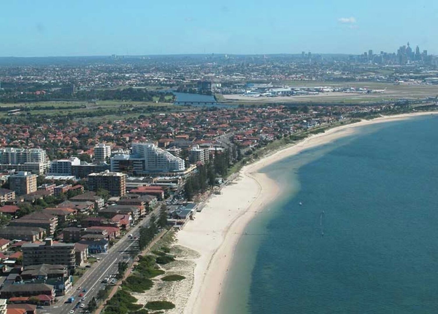 An aerial view of a coastal city with a wide beach, dense urban area, and a distant city skyline.