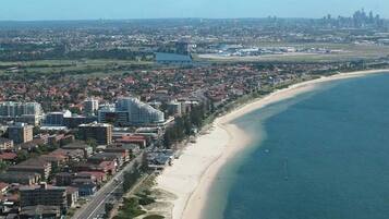 An aerial view of a coastal city with a wide beach, dense urban area, and a distant city skyline.