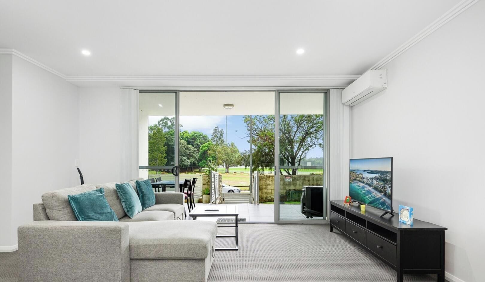 A modern living room with a grey sofa, teal cushions, a flat-screen TV, and a sliding glass door leading to an outdoor area.