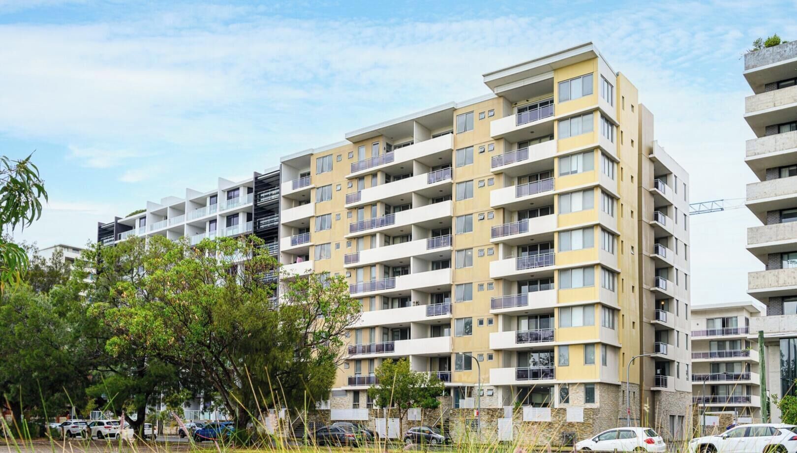 A multi-story residential building with balconies, a parking area, and surrounding greenery.