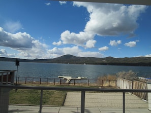 Blick auf einen See mit einer Holzterrasse, einem Metallgeländer und einem fernen Gebirge.