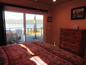 Ein Schlafzimmer mit Bett, Schminktisch, Stuhl und Blick auf einen See und Berge durch eine Schiebetür.