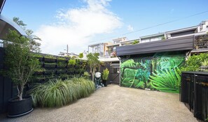 Un garage attenant à une maison, avec une porte de garage verte, une allée gravillonnée et une variété de plantes et d’arbres en pot.