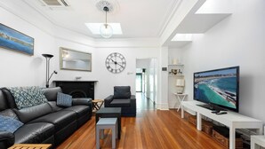 A living room with a fireplace, a large clock, a television, and a view of the beach. - KozyGuru | Glebe | Chic Stay - Steps to Light Rail (Glebe)