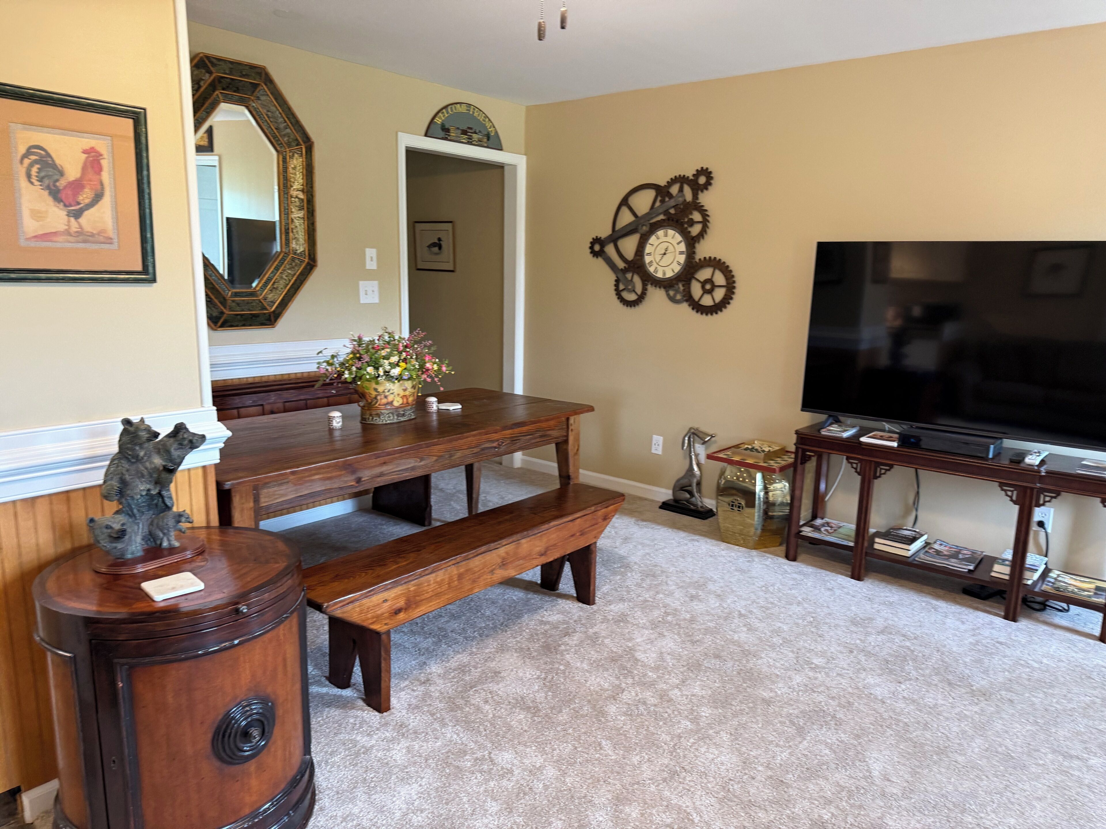A living room with a wooden dining table, benches, a TV stand with books, a clock, and a mirror.
