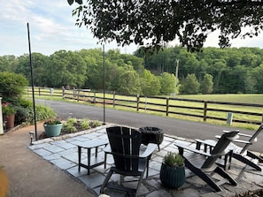 An outdoor seating area with chairs and tables, a paved walkway, and a view of a fenced field and trees.